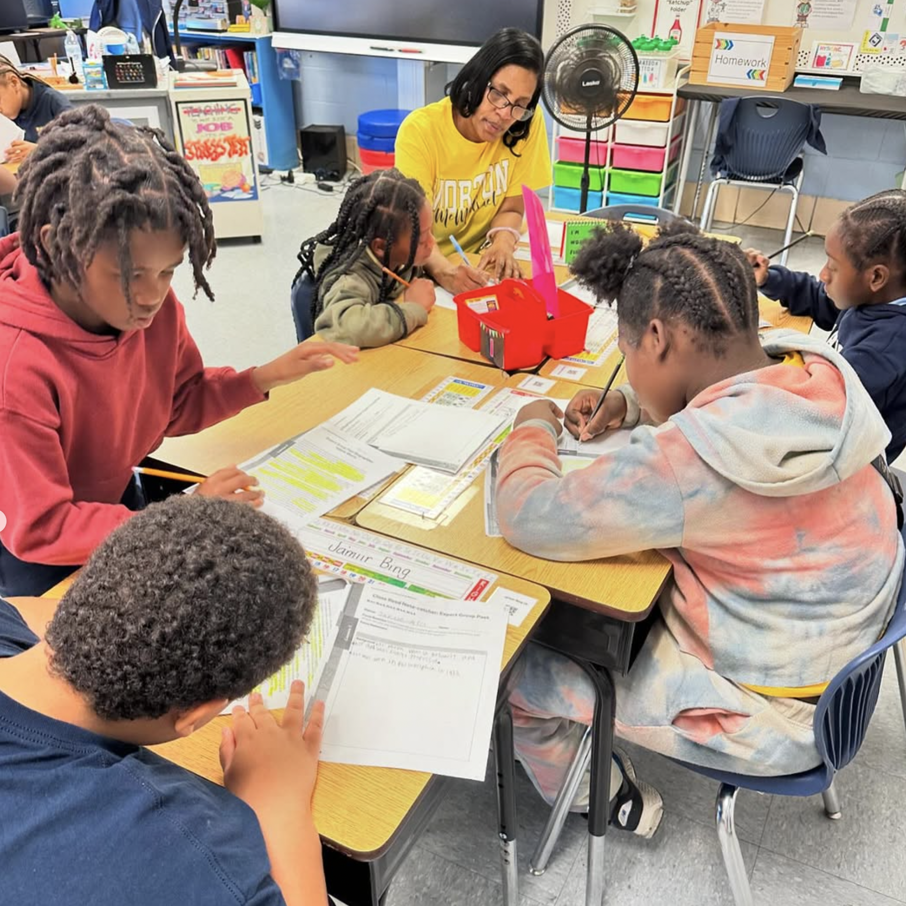 Students at Morton McMichael School sit around a table working on assignments with a teacher providing small group instruction in a classroom setting.